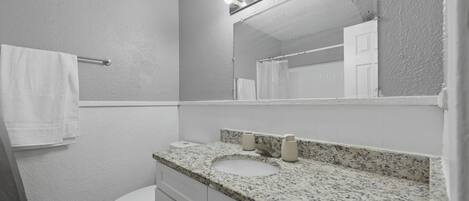 A bathroom with a white toilet, a granite countertop sink, a mirror with lights, and a towel rack.