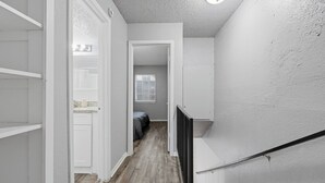 Ein schmaler Flur mit Holzboden, weißen Wänden und einer Tür zu einem Schlafzimmer mit einem Bett.