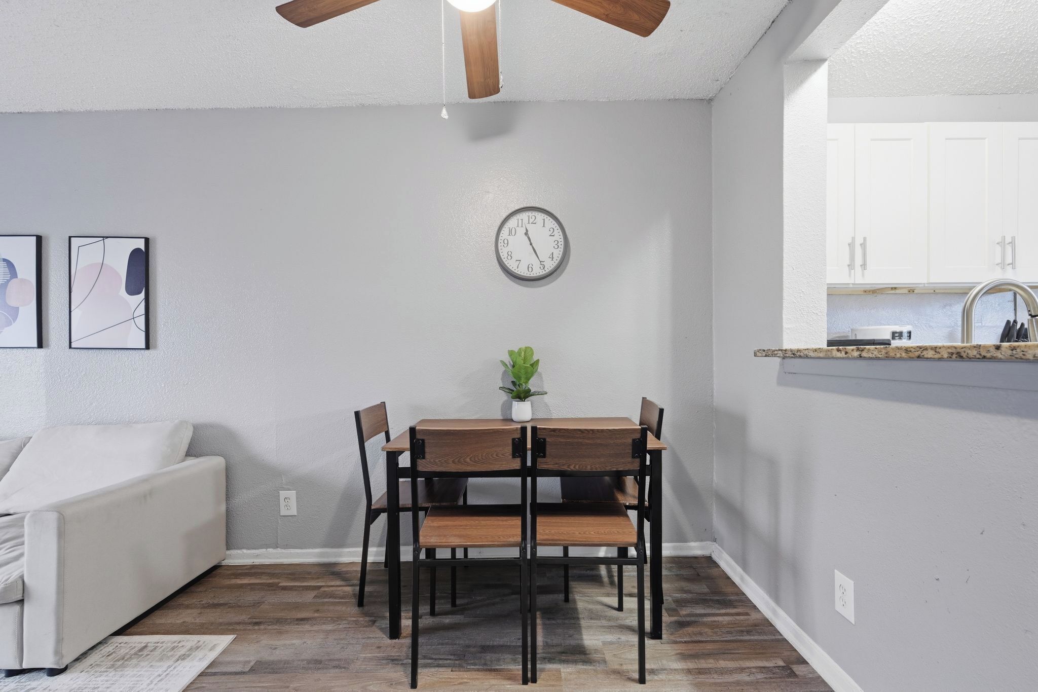 A dining area with a wooden table and chairs, a clock on the wall, a small plant, and a white couch in the background.