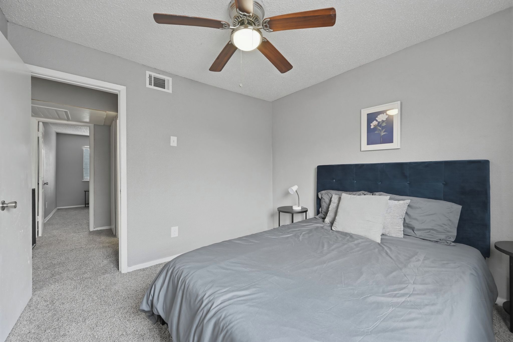 A bedroom with a bed, a ceiling fan, a bedside table, a nightstand, and a door leading to another room.