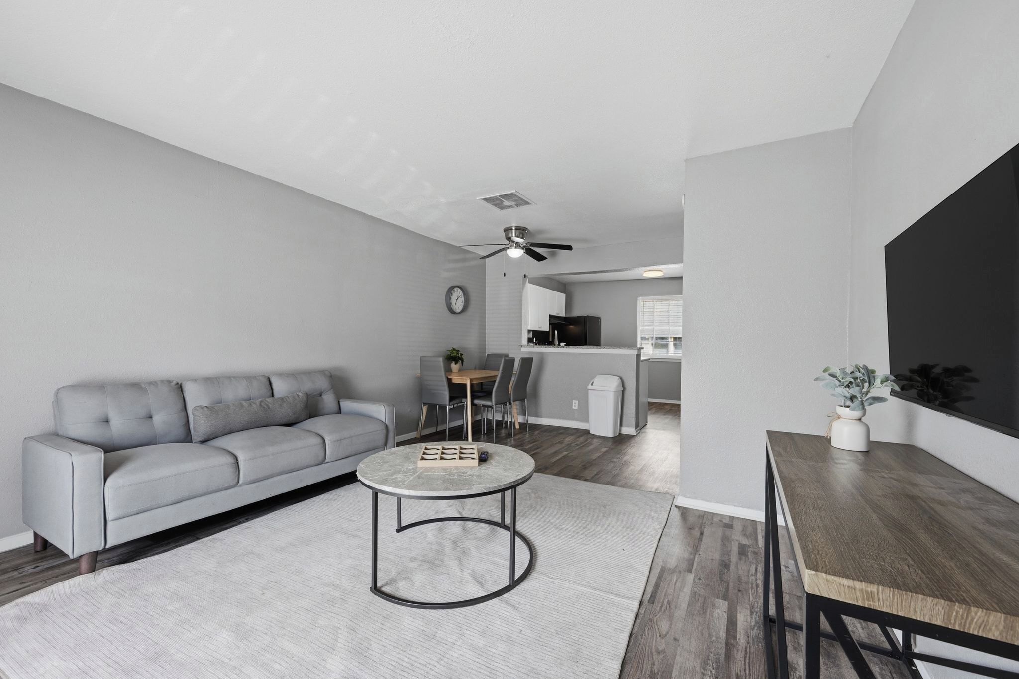 A modern living room with a grey sofa, a round coffee table, a dining table, and a kitchen area in the background.