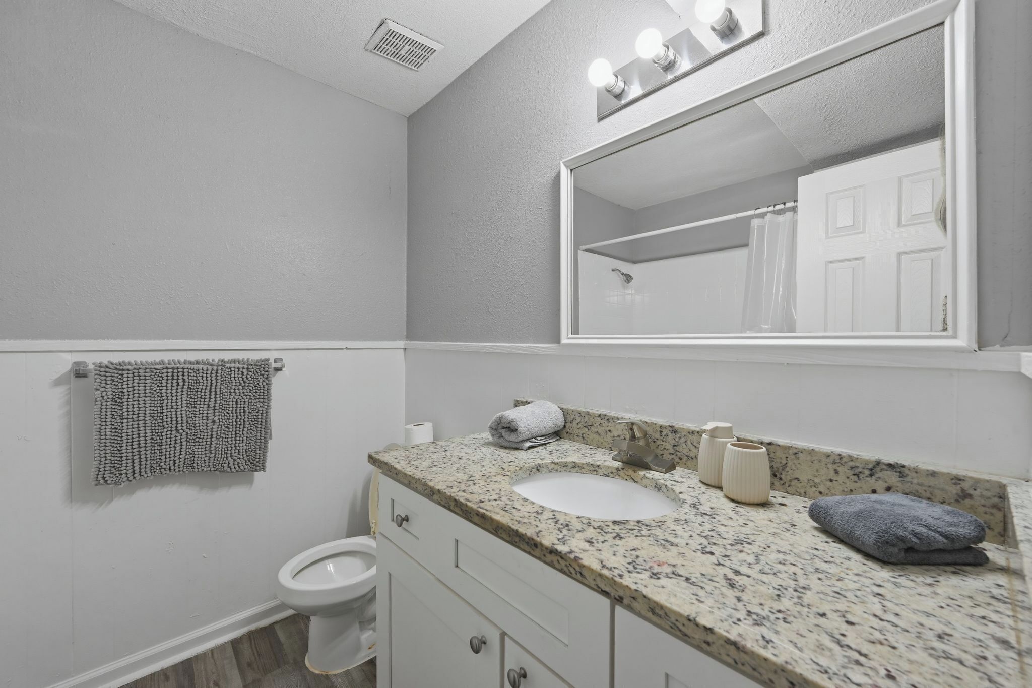 A bathroom with a white toilet, a sink with a granite countertop, a mirror with lights, and a towel rack.