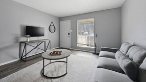 Een moderne woonkamer met een grijze bank, een ronde salontafel, een televisie op een zwart dressoir en een raam met jaloezieën.