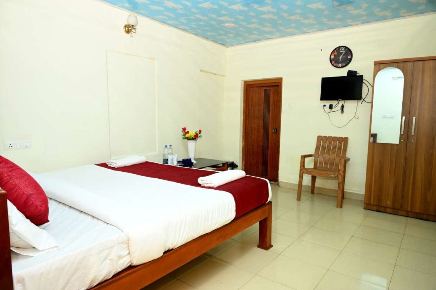 A hotel room with a bed, a chair, a TV, a clock, and a wooden wardrobe.