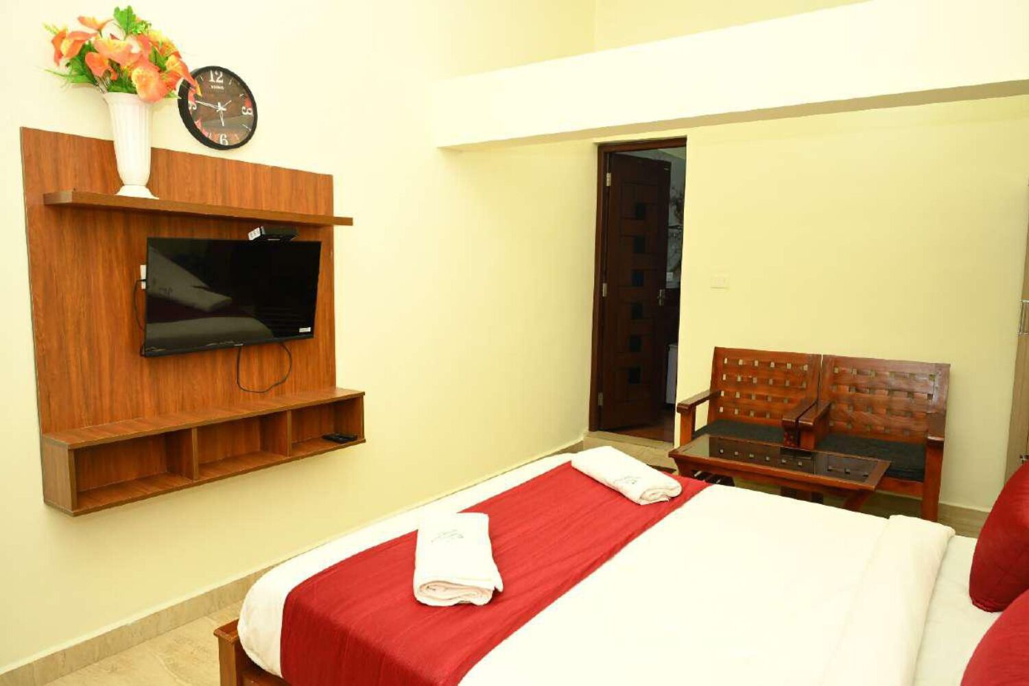 Quarto de hotel com uma cama, televisão presa a um painel de madeira, relógio e porta que dá para outro aposento.