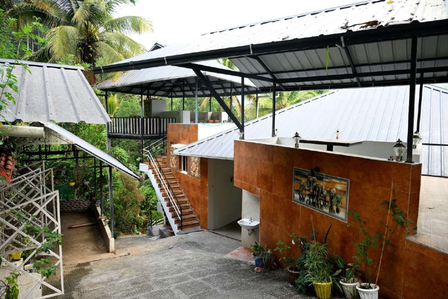 A covered outdoor area with a staircase leading to a second level, surrounded by potted plants and a metal roof structure.