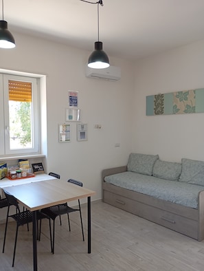 A room with a dining table, chairs, a sofa, a window with blinds, and a wall-mounted air conditioner. - Cozy open space with beautiful veranda (Castellana Grotte)
