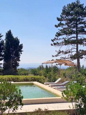 Una piscina con sillones de descanso y una pérgola, rodeada de árboles y vistas al paisaje.
