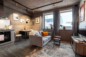 Ein modernes Wohnzimmer mit grauem Sofa, einem kleinen Esstisch, einer Küchenzeile mit Herd und großen Fenstern mit Vorhängen.