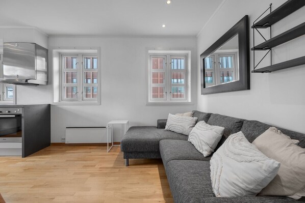 Un salon moderne avec un canapé gris, des coussins blancs, une petite table d’appoint blanche et un four encastré.