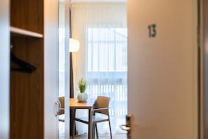 Une pièce avec une table en bois, deux chaises, une plante en pot et une fenêtre avec des stores.