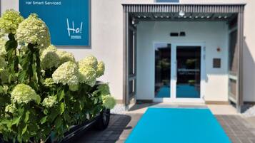 A modern building entrance with a blue carpet leading to a door, a sign with text, and a potted plant with white flowers.