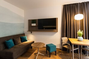 A modern living room with a sofa, a coffee table, a TV mounted on the wall, and a dining area with a table and chairs. - Harabruck Apartments (Gmünd)