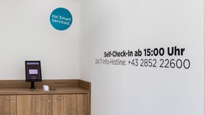 A self-check-in kiosk and a wall sign indicating self-check-in availability at 15:00 Uhr with a 24/7 info hotline number.
