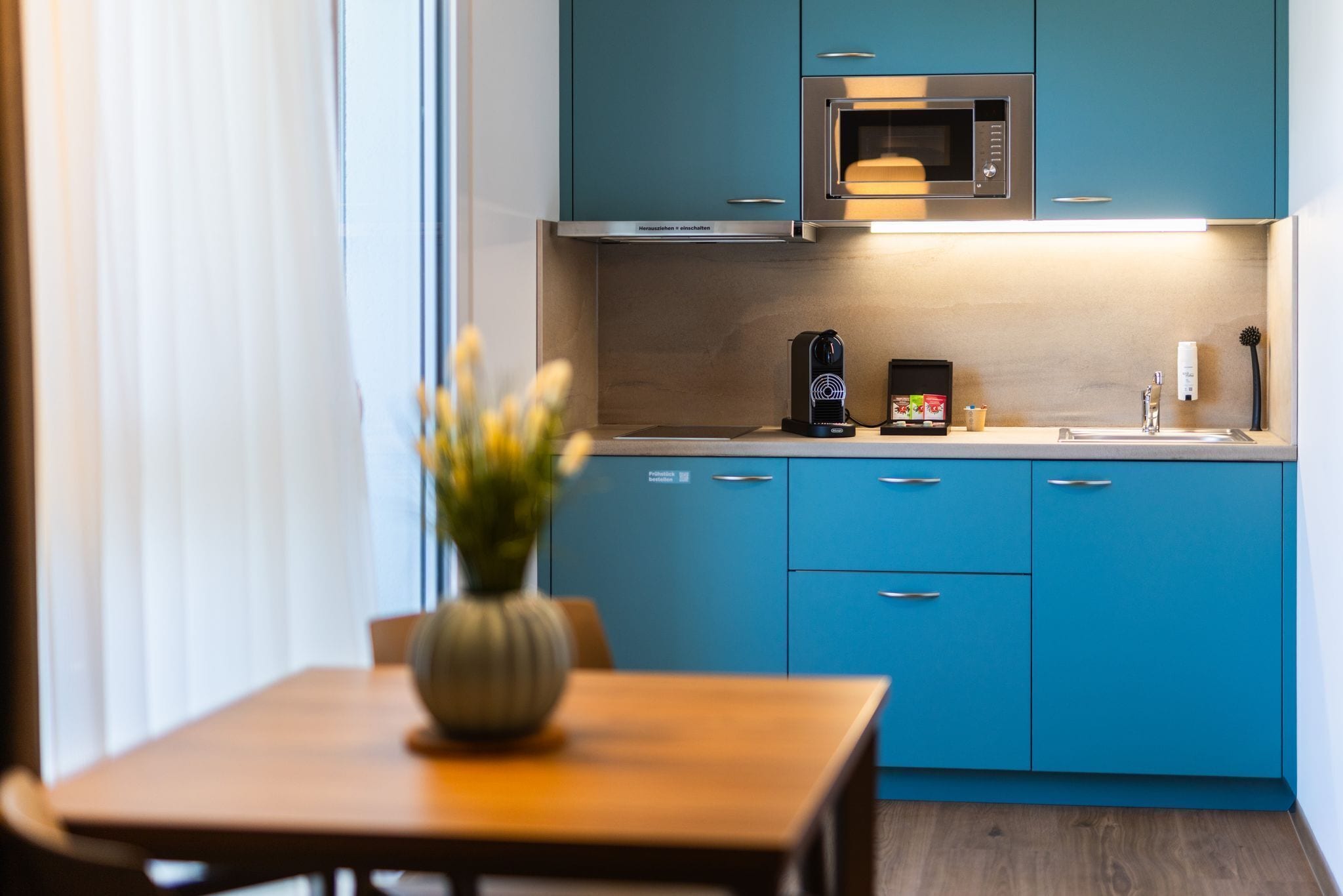 Eine moderne Küche mit blauen Schränken, einer Mikrowelle, einer Kaffeemaschine und einem Blumenstrauß auf dem Tisch.