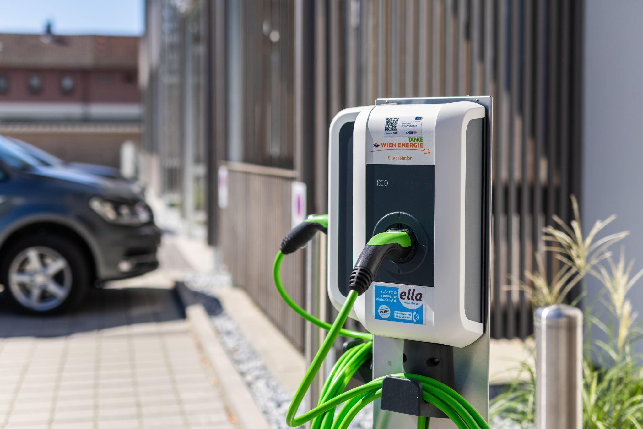 Una estación de carga para vehículos eléctricos con un código QR, un rótulo que dice 'Take Energie E-Ladestation' y un cable verde conectado a un coche.