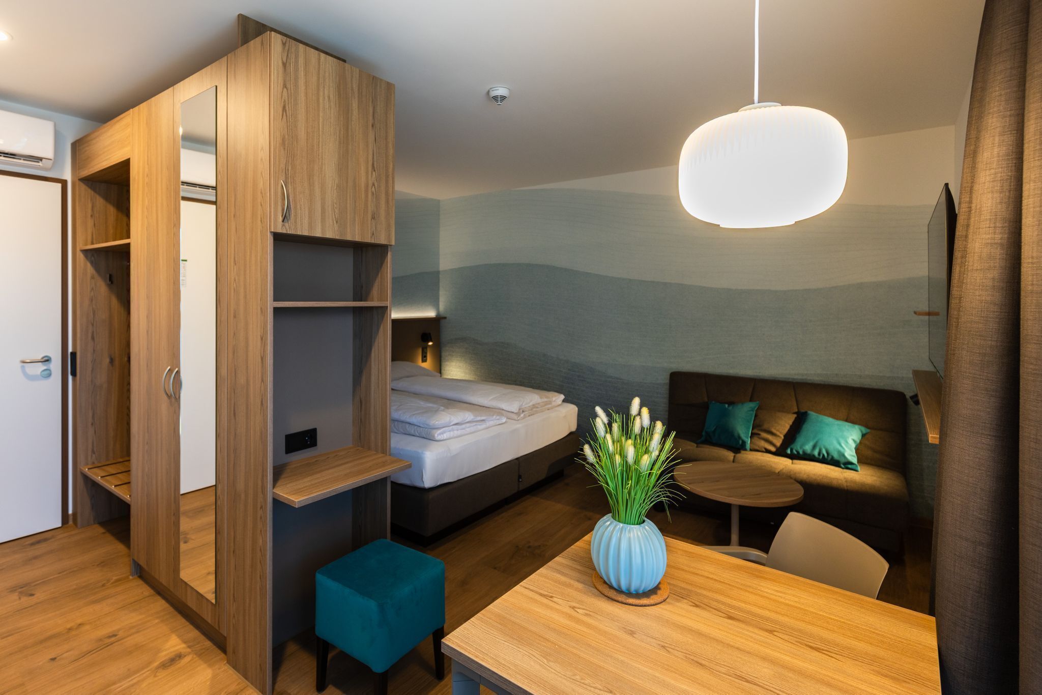 Une chambre d’hôtel compacte comprenant un lit, un coin salon avec un canapé et des coussins, une petite table et une armoire avec des étagères.