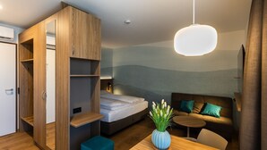 A compact hotel room with a bed, a seating area with a sofa and cushions, a small table, and a wardrobe with shelves.