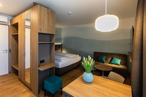 Een compacte hotelkamer met een bed, een zithoek met een bank en kussens, een klein tafeltje en een garderobe met planken.