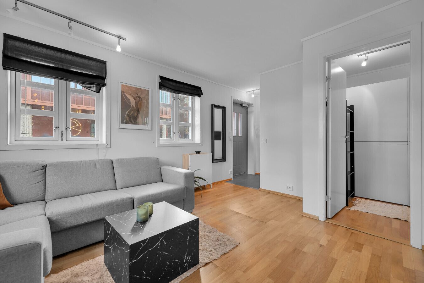 Een moderne woonkamer met een grijze bank, een marmeren salontafel en een houten vloer. Er zijn grote ramen met zwarte jaloezieën, een deur die leidt naar een hal, en een witte muur met een lijstwerkfoto.