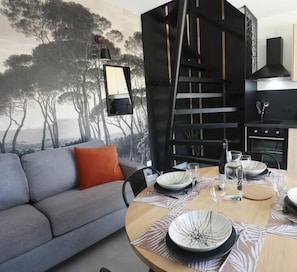 Uma sala de estar moderna com um sofá cinzento, uma mesa de jantar posta com pratos e copos, uma estante preta e um mural com árvores.