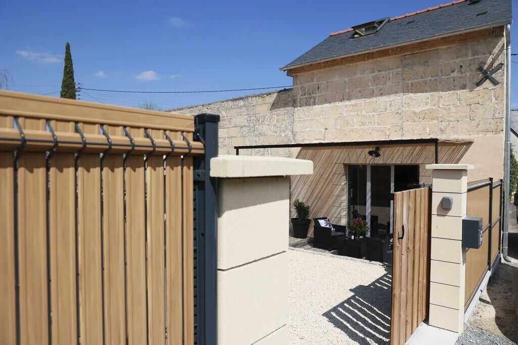 Une entrée fermée par un portail donnant accès à un immeuble à la façade en pierre, avec un portail en bois et un petit patio aménagé avec du mobilier d’extérieur.