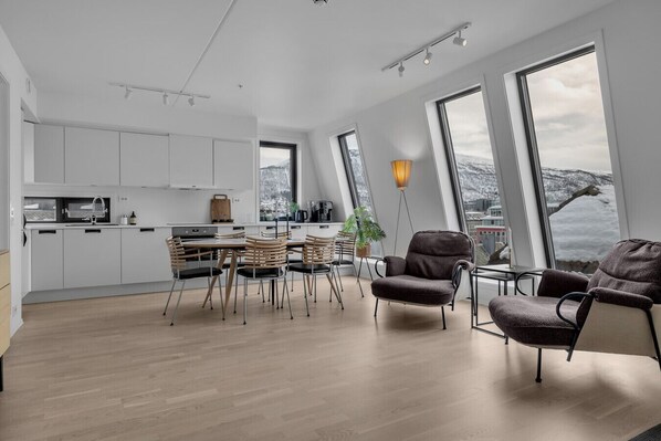 A modern kitchen with white cabinets, a dining area with wooden chairs, and large windows offering a view of a snowy landscape.