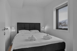 A minimalist bedroom with a bed, two wall-mounted lights, a window with a view of snow-covered hills.