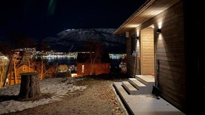 Eine Holzhütte mit Veranda, Baumstumpf und Blick auf eine beleuchtete Stadt und Berge im Hintergrund.
