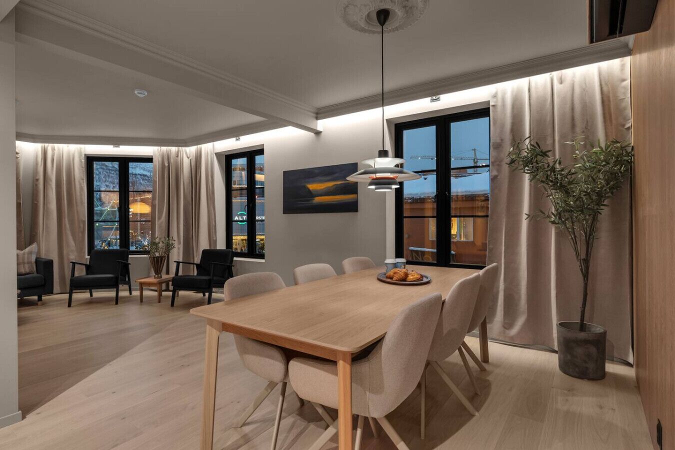 Un'area pranzo moderna con tavolo in legno, sei sedie, una lampada a sospensione e vista sulla città attraverso ampie finestre.