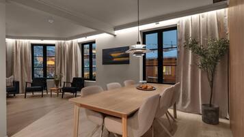 Un'area pranzo moderna con tavolo in legno, sei sedie, una lampada a sospensione e vista sulla città attraverso ampie finestre.
