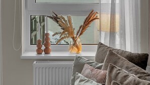 Ein Zimmer mit Blick auf eine Stadtlandschaft durch ein Fenster, ein Heizkörper darunter und eine Couch mit Kissen.