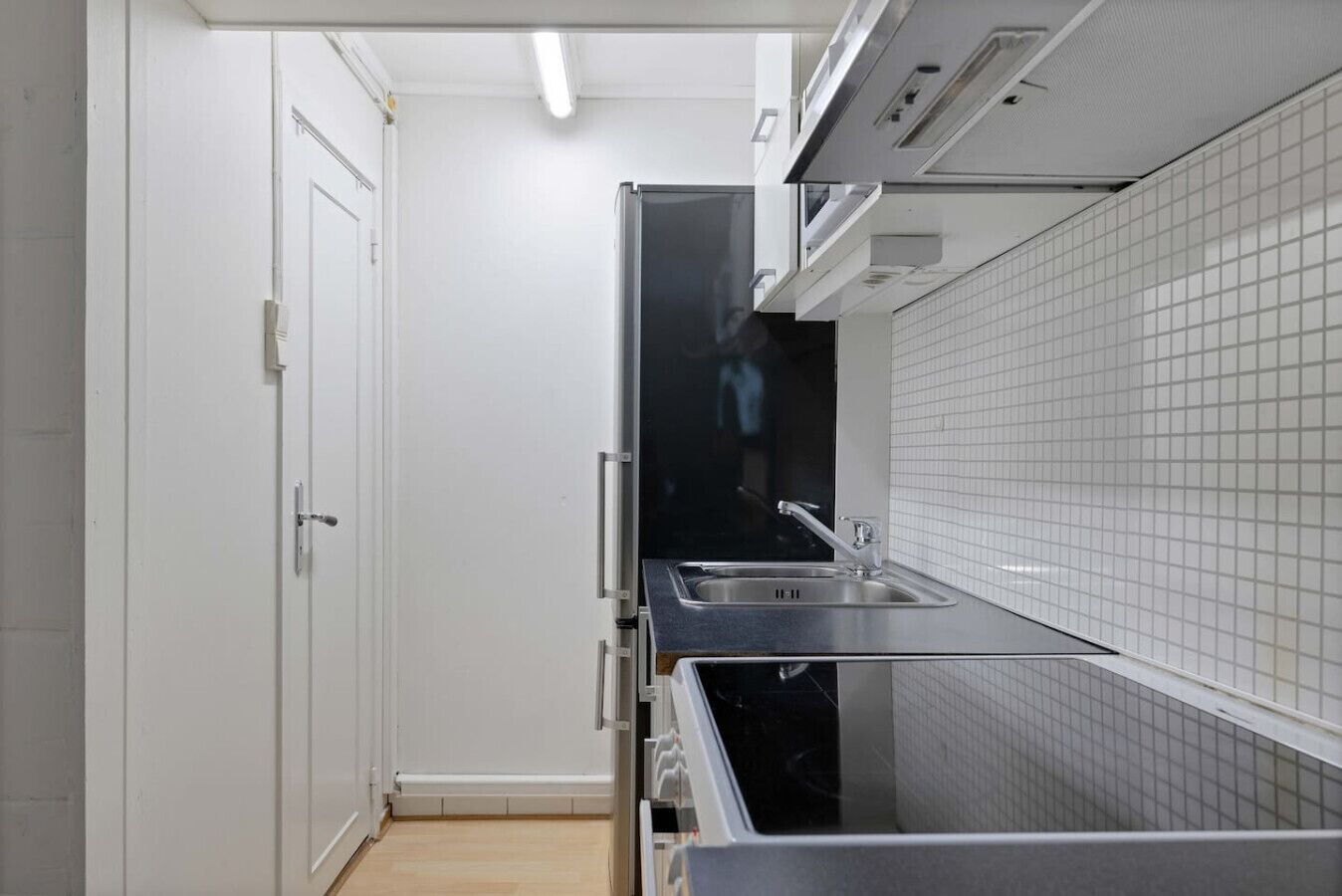 Une cuisine étroite avec un dosseret de carreaux blancs, des appareils électroménagers en acier inoxydable et une porte donnant sur une autre pièce.