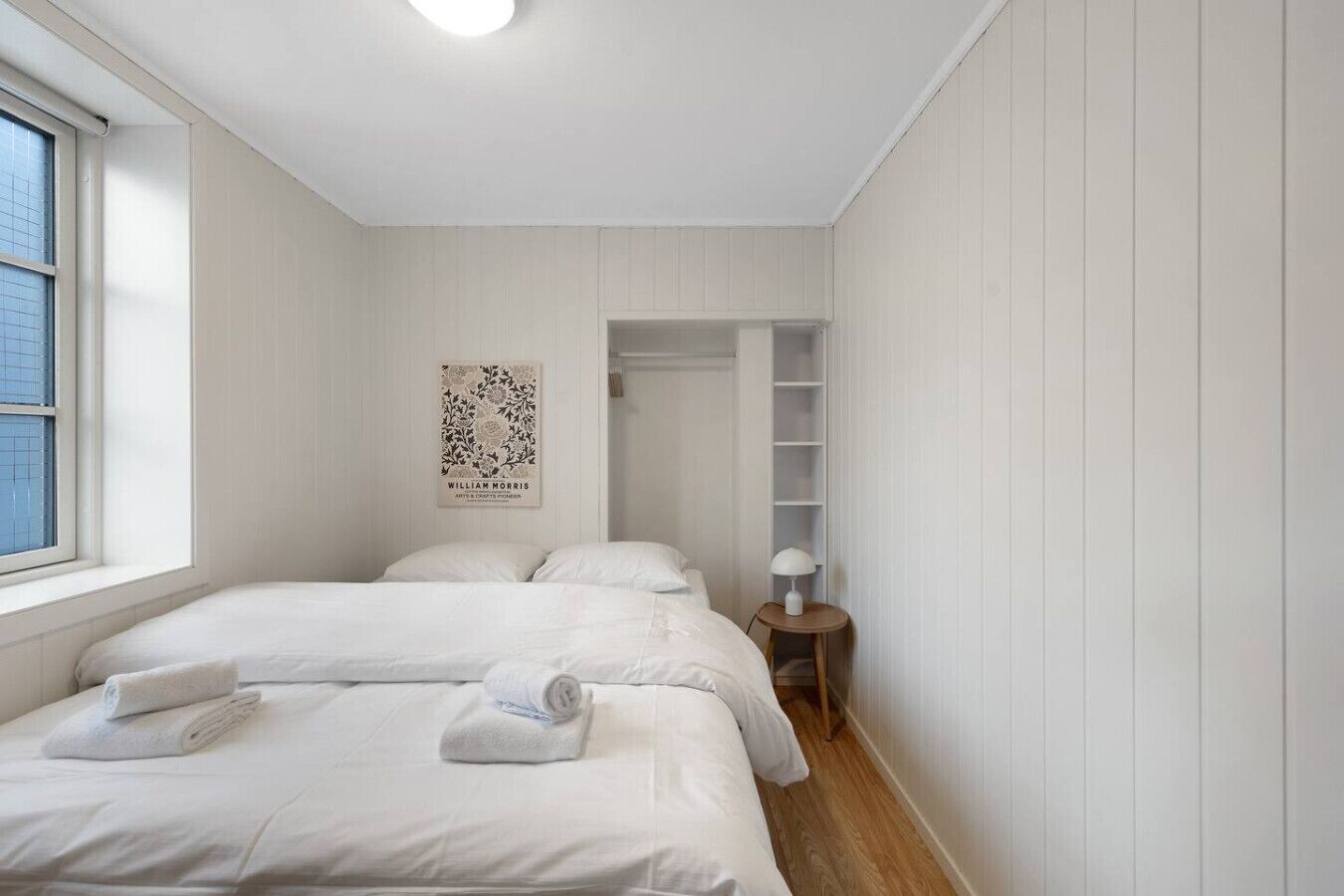 Une chambre avec deux lits, une fenêtre, une affiche accrochée au mur et une petite table de chevet avec une lampe.