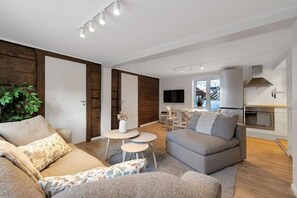 A modern living room with a sofa, a coffee table, and a TV. There is a kitchen area with a stove and cabinets.