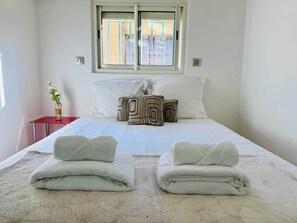 Un lit bien fait, avec des oreillers, une fenêtre avec des stores, une petite table rouge sur laquelle est posé un vase, et un interrupteur fixé au mur.