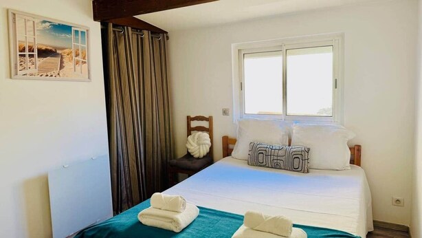 A neatly made bed with white linens, a teal bedspread, and two folded towels. There is a window with white curtains, a wooden nightstand with a lamp, and a framed picture on the wall. - "La Plagette" Baraquette near Etang, Clim (Sète)
