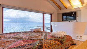 A bedroom with a large window overlooking the sea, a bed with a colorful quilt, a small bathroom with a mirror and sink, and a television on the wall. - Almoxarife Beach House (Horta)