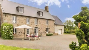 Un bâtiment en pierre avec une allée pavée, un espace extérieur aménagé avec une zone de repos couverte et un jardin.