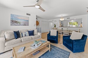 Un salon avec un canapé beige, un fauteuil bleu, une table basse en bois et un coin repas comprenant une table et des chaises en bois.