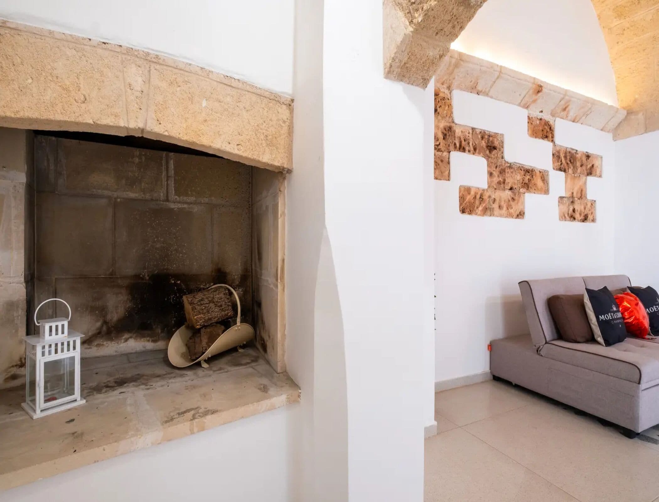 Una chimenea con detalles de piedra, un farol blanco y un sofá gris con cojines.