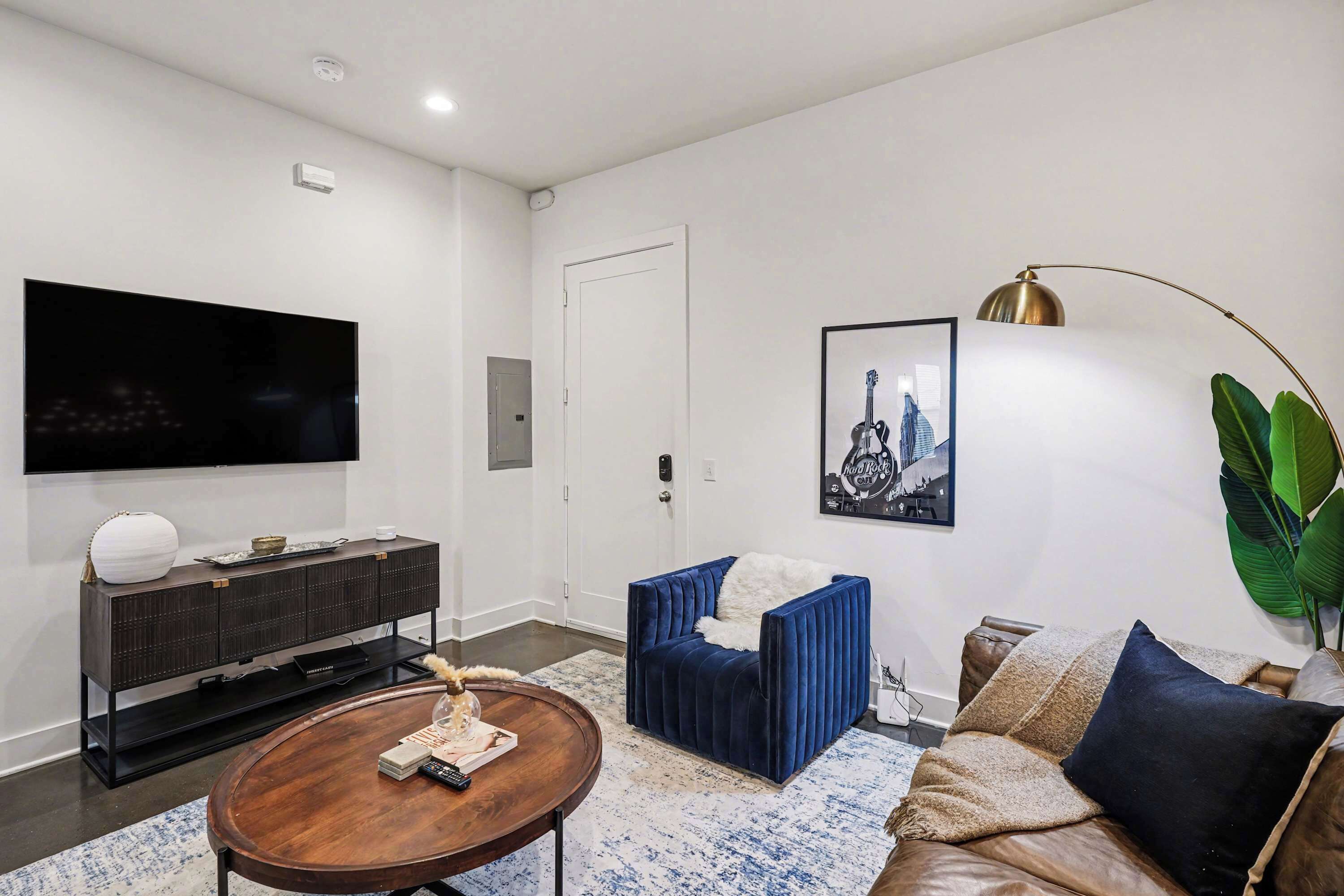 Een moderne woonkamer met een flatscreen televisie, een tv-meubel van donker hout, een blauwe fluweel fauteuil, een bruine leren bank en een grote groene plant.