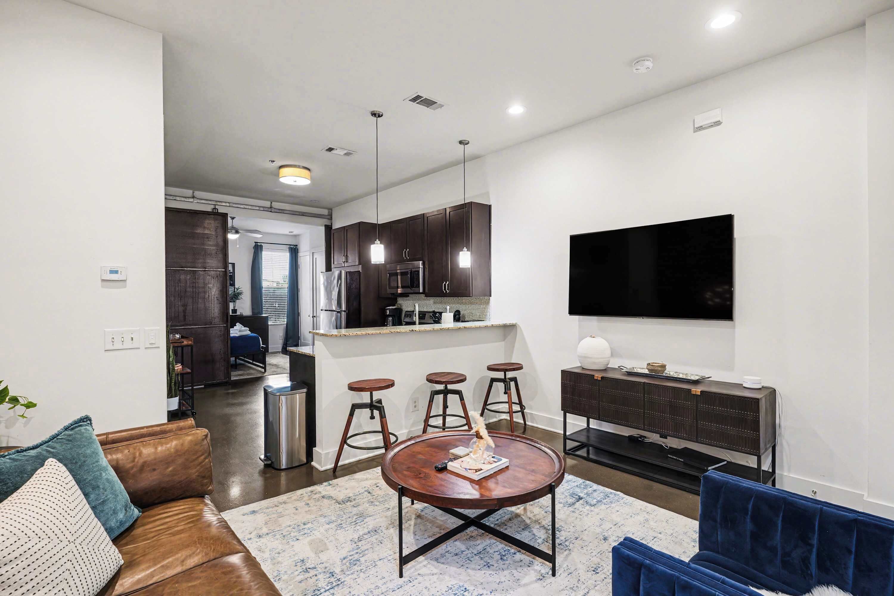 A modern living room with a brown leather sofa, a round coffee table, and a blue velvet sofa. There is a kitchen area with dark cabinets and a flat-screen TV mounted on the wall.