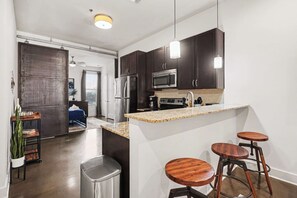 A modern kitchen with dark cabinets, a stainless steel refrigerator, a microwave, and a sink. There are three wooden stools at the island. - Trendy Condo for 2 near Centennial park & Vandy! (Nashville)