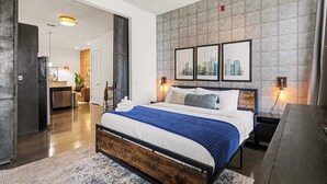 Et moderne hotelværelse med en stor seng, et træhovedgærde, bykunst på væggen, et skrivebord og et badeværelse, der kan ses gennem en åben dør.