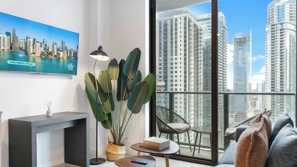 A modern living room with a cityscape view, a large plant, a sofa, a side table, and a console table. - Urban 1BR+Den | Gym | Spa | Downtown Miami (Miami)