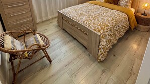 Una camera da letto con un letto, una lampada da comodino, una sedia in vimini, una cassettiera in legno e una finestra con tende.
