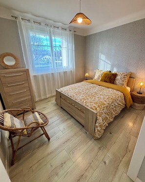 Ein Schlafzimmer mit einem Bett, einer Nachttischlampe, einem Korbstuhl, einer Holzkommode und einem Fenster mit Vorhängen.