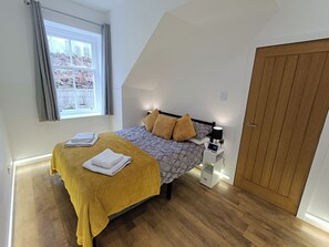 A bedroom with a bed, a nightstand, a wooden door, and a window with curtains. - RnB - The Coach House - Ev Charger Option (Girvan)