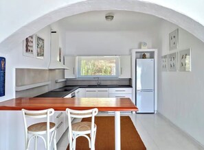 A compact kitchen with white cabinetry, a wooden table, two wicker chairs, and a window providing natural light. - Sunset Vibes villa (Mykonos)